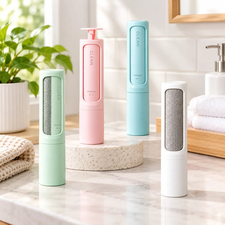Four CLEAN pet hair remover tools in mint green, pink, light blue, and white are arranged on a bathroom counter with a stone pedestal, a potted plant, and folded towels in the background.
